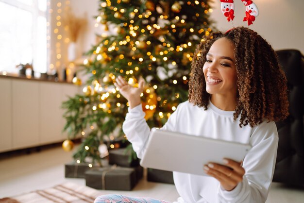 A young woman on the background of a Christmas tree with gifts with a tablet has a video call.