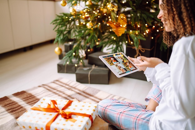 A young woman on the background of a Christmas tree with gifts with a tablet has a video call.