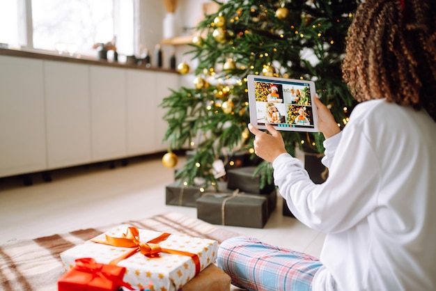 A young woman on the background of a Christmas tree with gifts with a tablet has a video call.