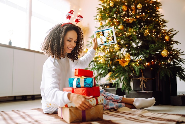 A young woman on the background of a Christmas tree with gifts with a tablet has a video call.