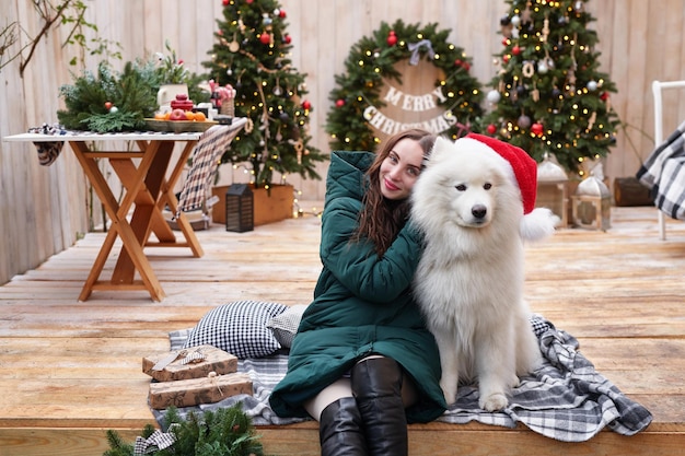 Young woman on background of Christmas tree with white samoyed dog in santa hat outdoors Yard decoration for New Year