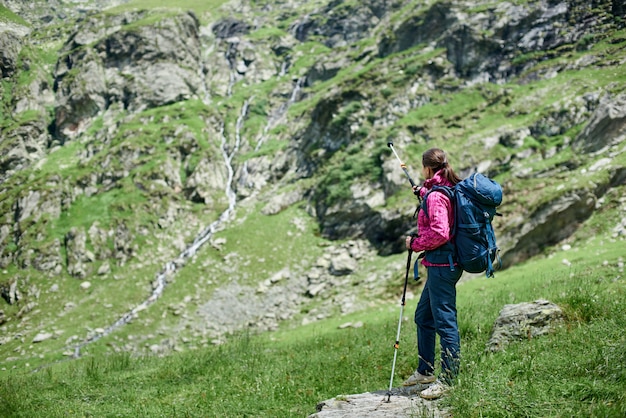 Young woman backpacker with trekking sticks and a backpack is standing on a stone