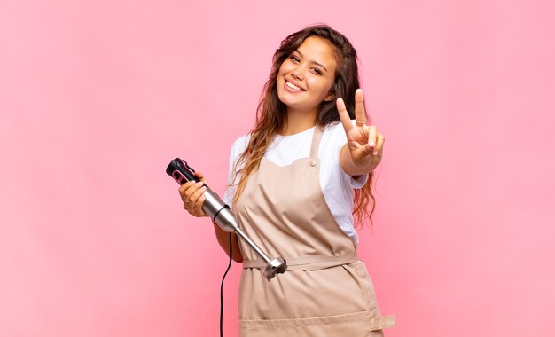 若い女性のパン屋は笑顔で幸せそうに見え、のんきで前向きで、片手で勝利または平和を身振りで示す