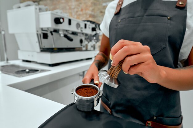 Young woman barista presses ground coffee using tamper in coffee shop