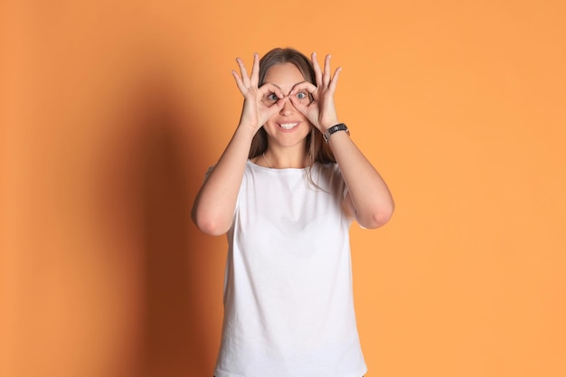 Young woman in basic clothing smiling looking at camera through holes made with fingers isolated over yellow background.