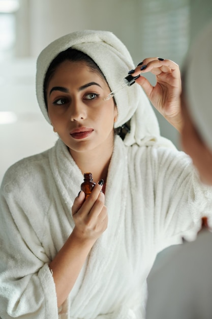 Young woman in bathrobe applying moisturizing serum on her face after shower