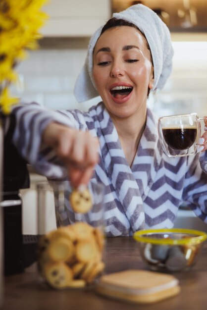 バスローブを着た若い女性が家でクッキーを食べ、朝のコーヒーを飲みます。