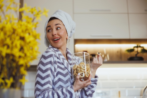 Young woman in bathrobe holding jar of cookies in her kitchen and enjoying morning at home.