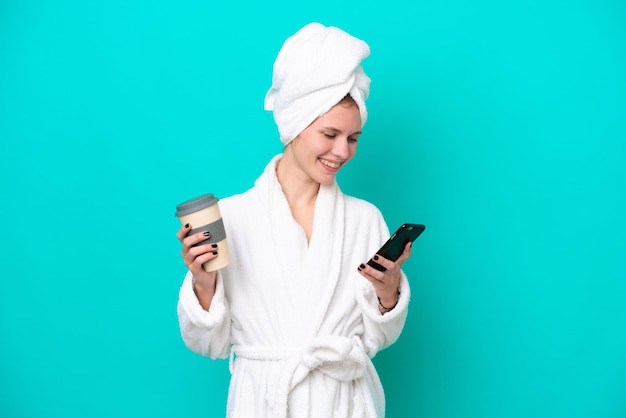 Young woman in a bathrobe isolated on blue background holding coffee to take away and a mobile