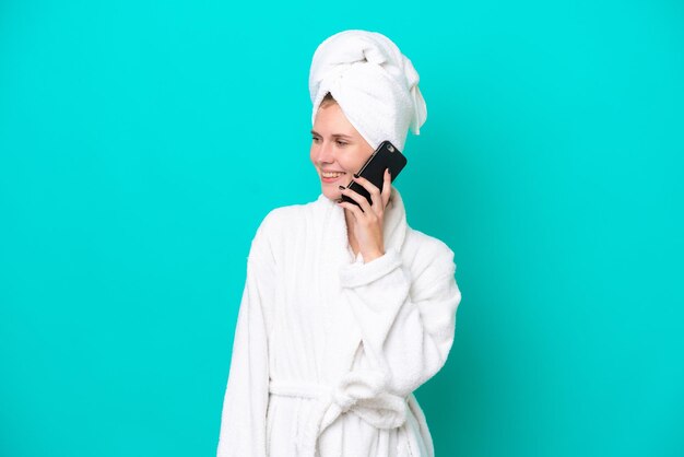 Young woman in a bathrobe isolated on blue background keeping a conversation with the mobile phone with someone