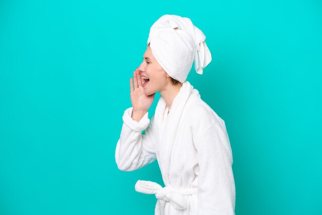 Young woman in a bathrobe isolated on blue background shouting with mouth wide open to the lateral