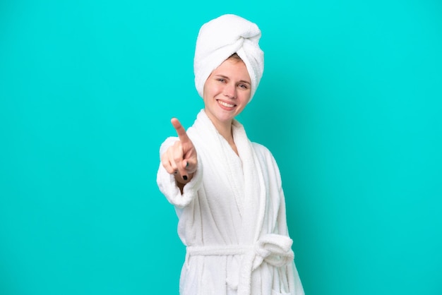 Young woman in a bathrobe isolated on blue background showing and lifting a finger