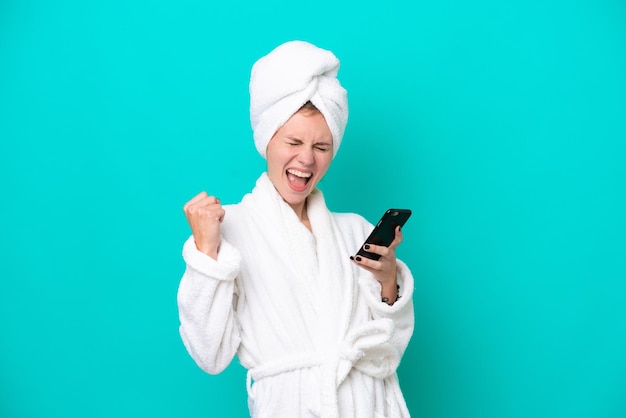 Young woman in a bathrobe isolated on blue background with phone in victory position