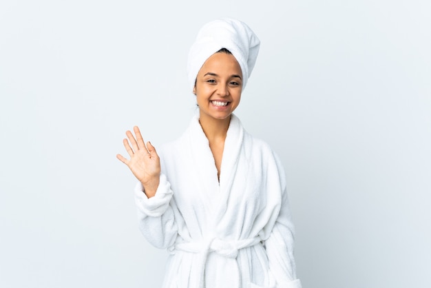 Young woman in bathrobe over isolated white wall saluting with hand with happy expression