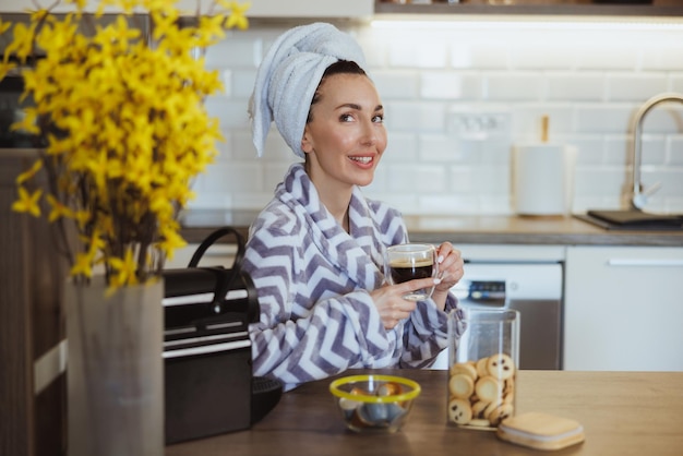 バスローブを着た若い女性が、自宅のキッチンでリラックスして朝のコーヒーを飲みます。