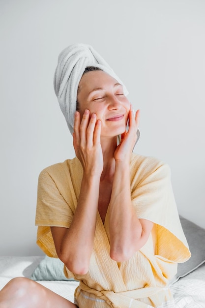 Young woman in a bathrobe and a towel on her head applies a cream to her skin massages her face