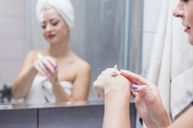 浴室の若い女性はタオルで美しさと健康を高めるために化粧品の手順を保持します