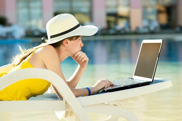 Young woman on beach chair at swimming pool working on computer laptop connected to wireless internet typing text on keys in summer resort