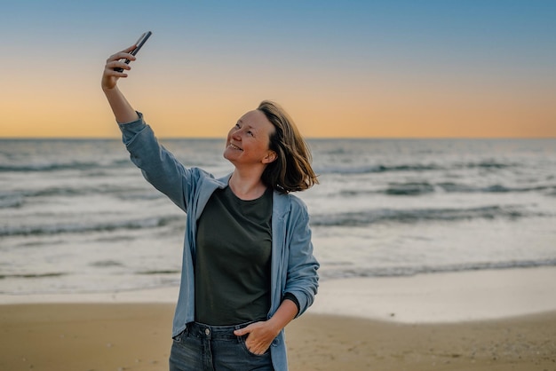 A young woman on the beach near the ocean in the spring at sunset takes a selfie talks via video