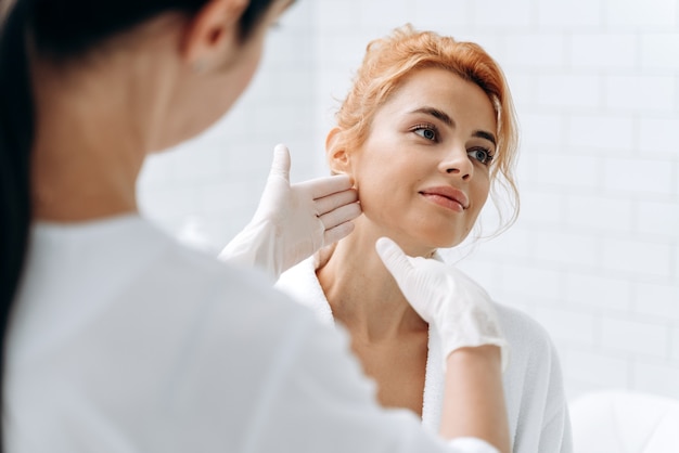 Young woman at beauty clinic cosmetology service sitting on medical chair while female doctor wearing gloves holding her face and examining skin