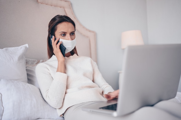 Young woman in bed, speaking phone and working on a laptop.