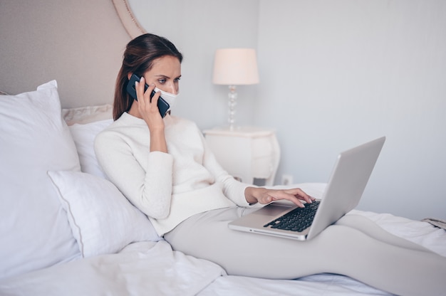 Молодая женщина в постели, говоря телефон и работает на ноутбуке.