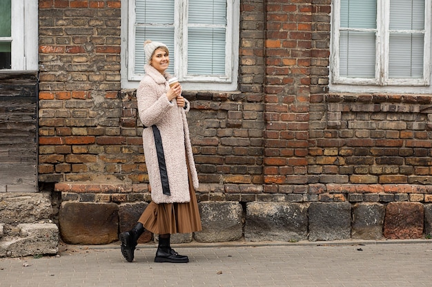 Young woman in beige faux fur hat, coat with disposable recyclable cup of coffee in hands walks through city in background of old type red brick building. smiles happily enjoying autumn day.