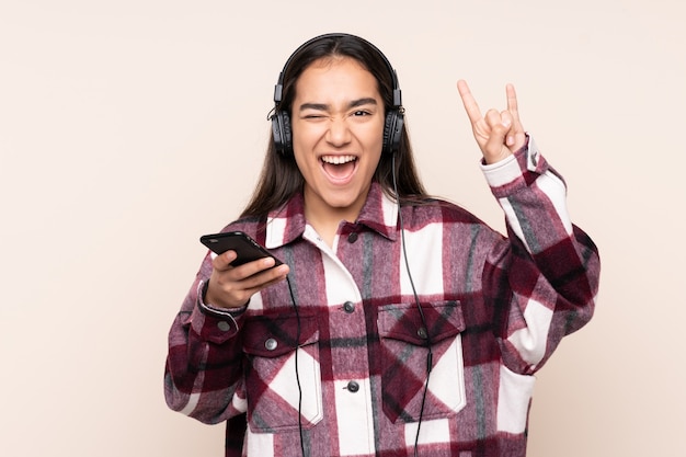 Young woman on beige listening music with a mobile making rock gesture