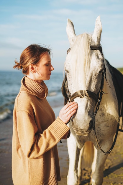 Молодая женщина в бежевом свитере с белой лошадью на фоне морского пейзажа