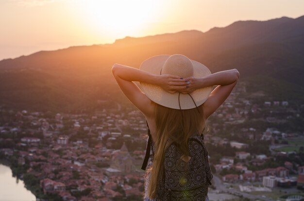 Young woman in big hat looking at sunset over the city and put hands on head