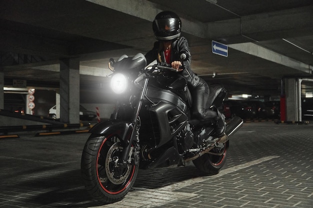 Motociclista della giovane donna in un casco che si siede su una moto nel garage sotterraneo