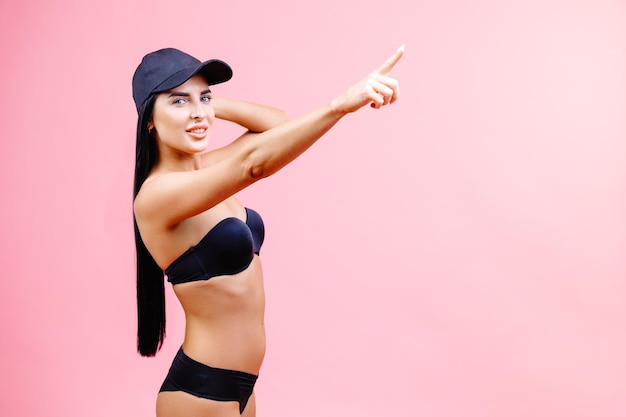 Young woman in bikini in summer holidays standing and pointing finger to the side on pink background in studio