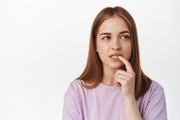 Young woman bite finger while ponder smth, looks away thoughtful, calculating in mind, makes decision, standing against white wall