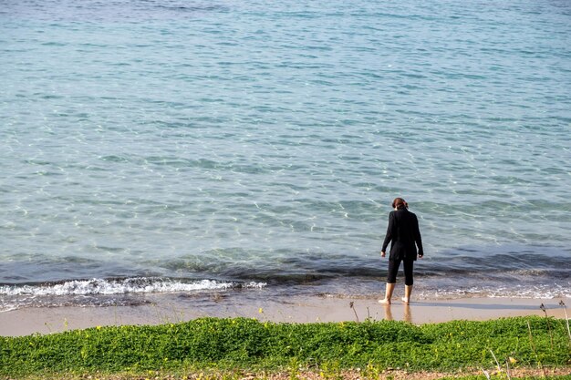 Young woman in black barefoot stands on Greek sandy beach waiting for the sea wave to wet her leg