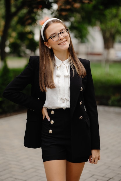 A young woman in a black blazer and glasses stands on a sidewalk in a park.