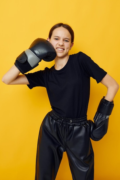 Молодая женщина в черных боксерских перчатках позирует на желтом фоне спорта