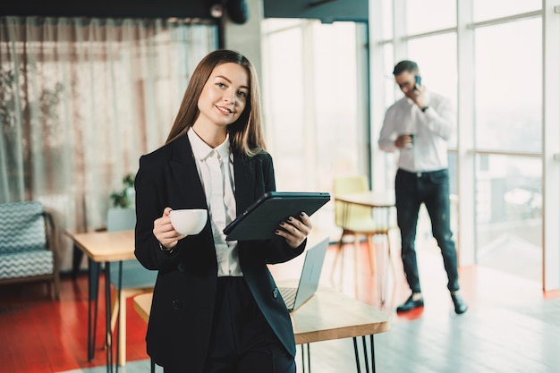검은색 클래식 슈트를 입은 젊은 여성이 현대적인 사무실에 서서 커피를 마시고 휴대용 태블릿에서 사무실에서 일하고 있습니다.