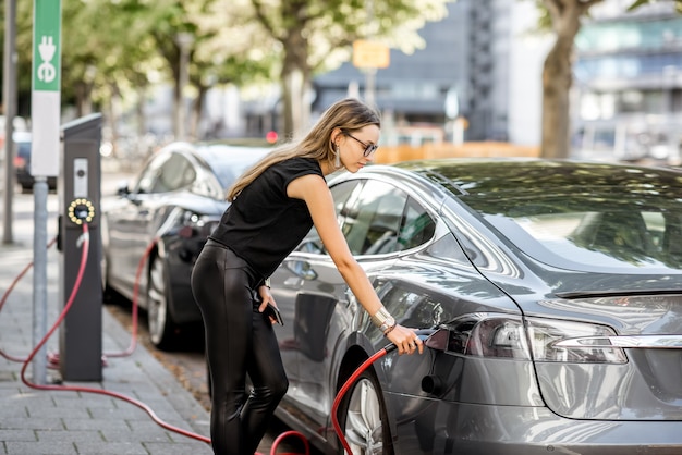 검은 옷을 입은 젊은 여성이 로테르담 시의 거리에서 야외 전기 자동차에 커넥터를 연결합니다.