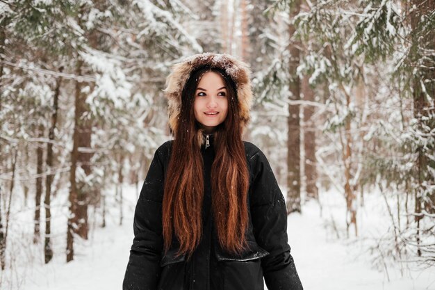 Foto giovane donna in cappotto nero che cammina nella foresta invernale e sorridente nel parco davanti agli alberi all'aperto