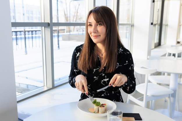 黒いドレスを着た若い女性が、スタイリッシュな街のカフェで昼食時にロールパンを食べる