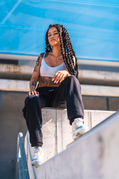 Young woman of black ethnicity with long braids and with tattoos sitting on the cement Urban shoot