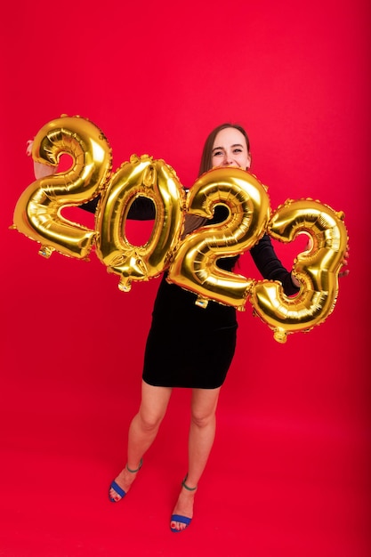 A young woman in a black evening elegant dress and highheeled shoes holds the numbers of the new year 2023 on a red background