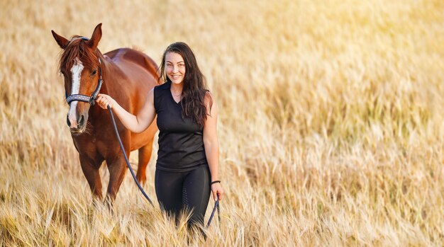 Young woman in black leggings and t shirt walking with brown Arabian horse in wheat field