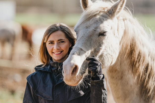 Young woman in black riding jacket standing near white Arabian horse smiling happy closeup detail