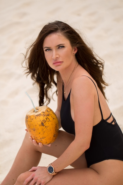 Молодая женщина в черном купальнике с кокосовым коктейлем на пляже