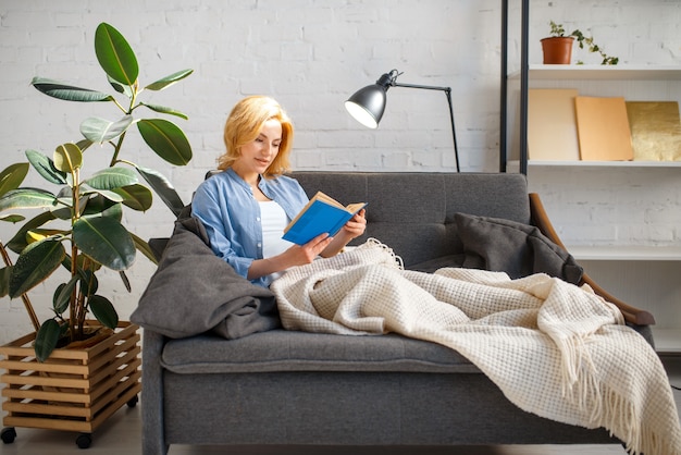 Young woman under a blanket reading a book on cozy yellow couch, living room in white tones