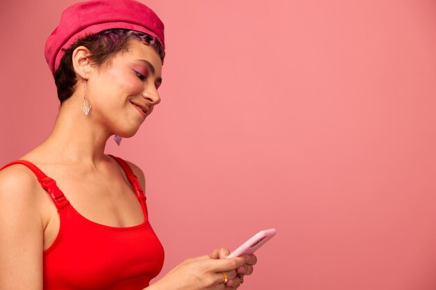 Young woman blogger with colored pink hair and short haircut flipping through the screen of the phone and typing a message with a smile in stylish clothes on a pink background