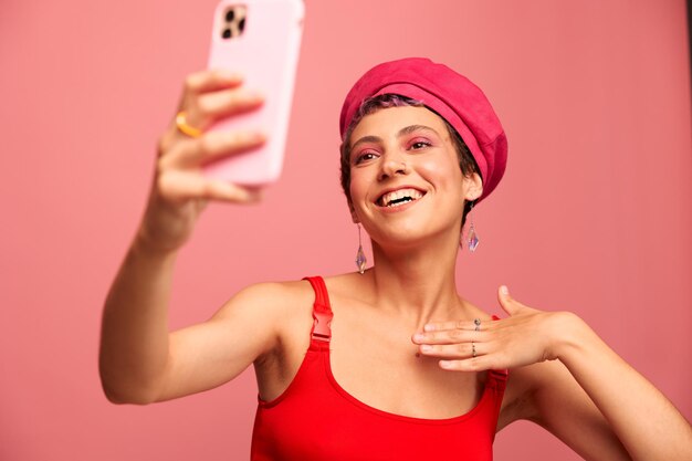 ピンク色の髪とショートヘアの若い女性ブロガーが電話で自分の写真を撮り、ピンクの背景のモノクロ スタイルにスタイリッシュな服と帽子で笑顔を放送します