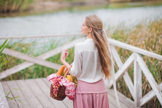 Young woman blonde with long hair in retro style vintage clothes stands with a picnic basket on a wooden pier of the lake