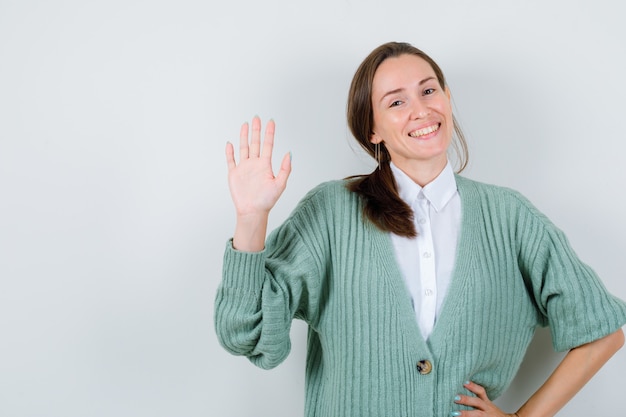 Young woman in blouse, cardigan greeting with open hand and looking jovial , front view.